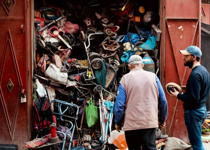 Two men inspecting a workshop with a cluttered collection of discarded items and tools.