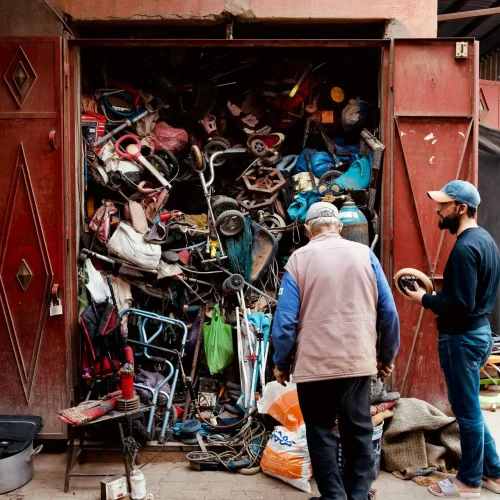 Two men inspecting a workshop with a cluttered collection of discarded items and tools.
